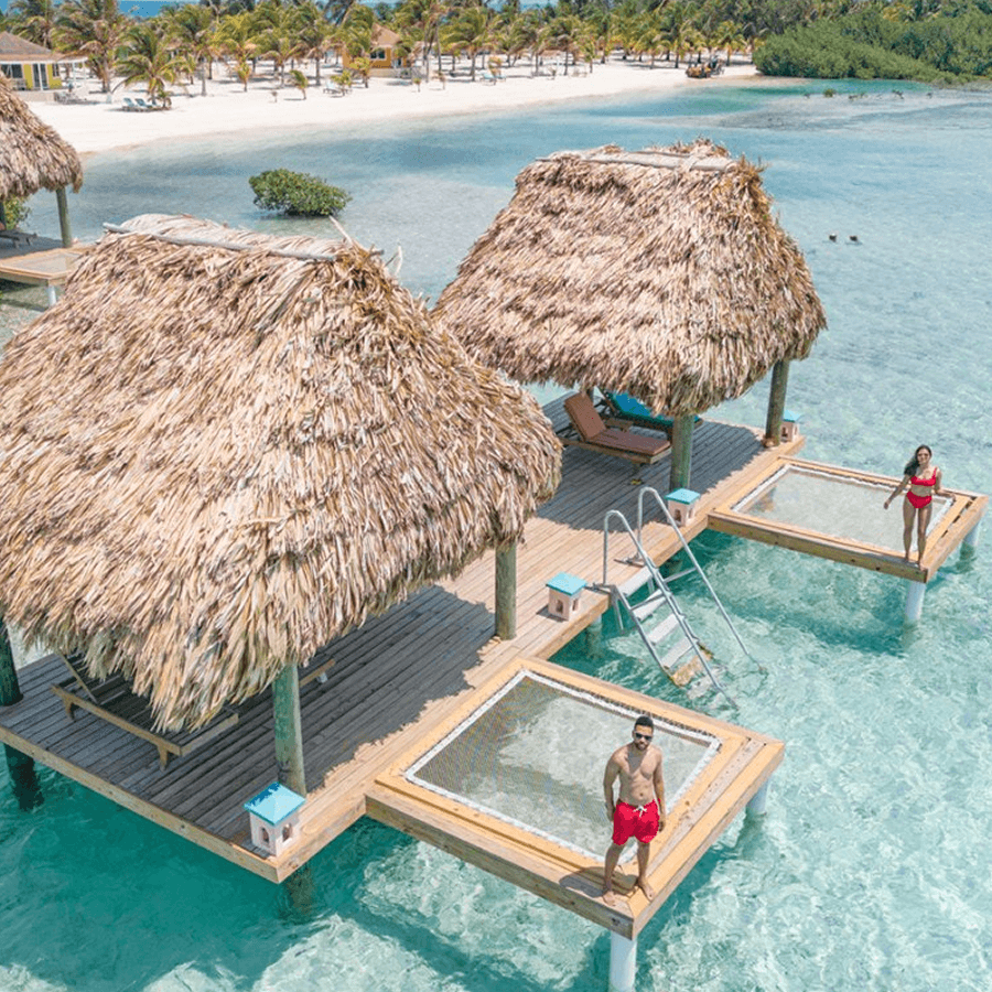 An aerial view of a luxurious overwater bungalow resort. The bungalows have thatched roofs and wooden decks, with glass floors looking down into the crystal-clear water. A couple is standing on one of the decks, enjoying the view.