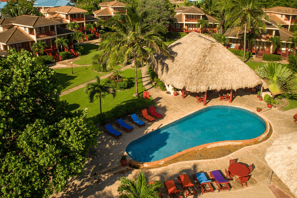 An aerial view of a tropical resort featuring a swimming pool surrounded by colorful lounge chairs and a thatched-roof bar. Several buildings with red roofs are visible, and palm trees dot the landscape.