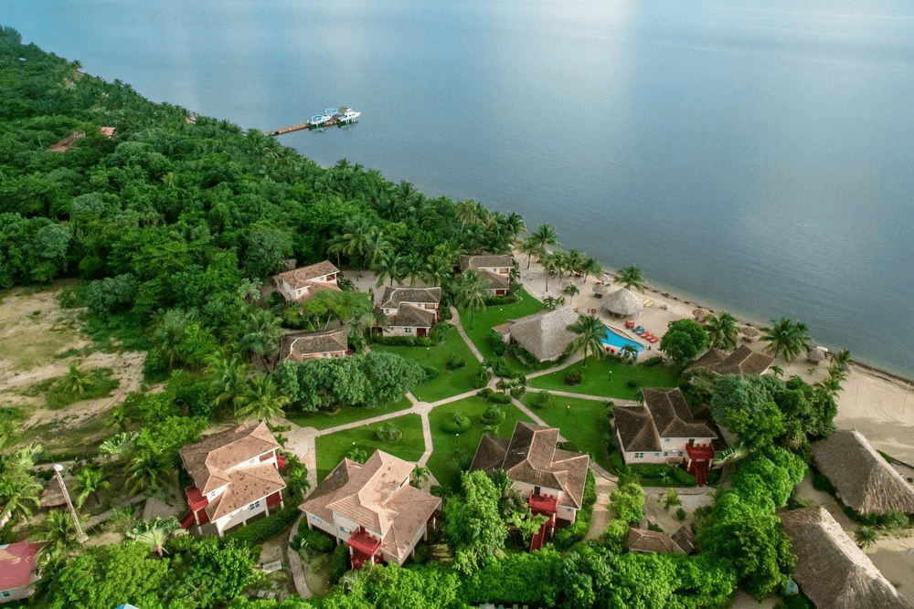 An aerial view of a beachfront resort in Belize. The resort features multiple buildings, a swimming pool, a pier, and lush green vegetation. The ocean and a nearby town are visible in the background.