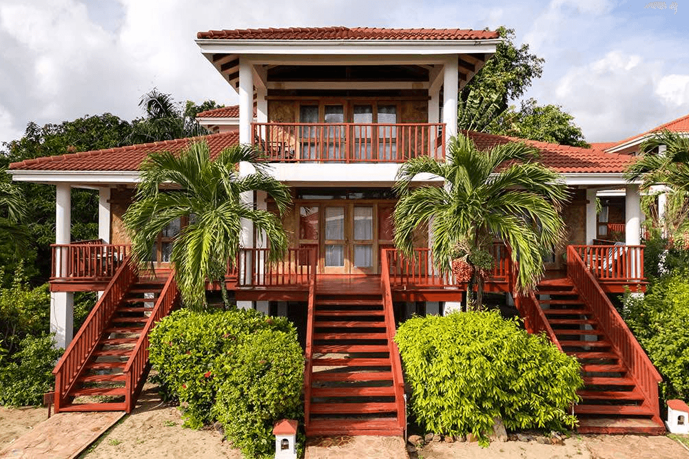 A two-story beachfront villa with a red tiled roof and white walls. The villa features large wooden decks and staircases, surrounded by palm trees and lush greenery.
