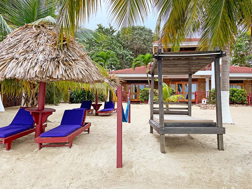 A sandy beach area featuring purple lounge chairs, a thatched umbrella, and a shaded structure surrounded by tropical greenery.