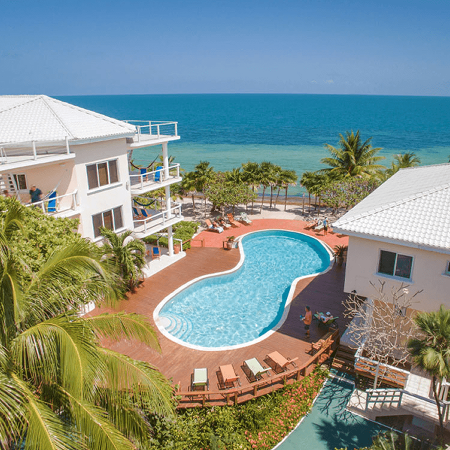 An aerial view of a beachfront resort with white buildings, a large swimming pool, and palm trees. The ocean is visible in the background.