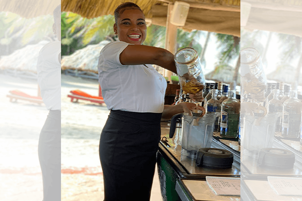 A smiling bartender makes a drink in a blender at an outdoor bar on the beach.