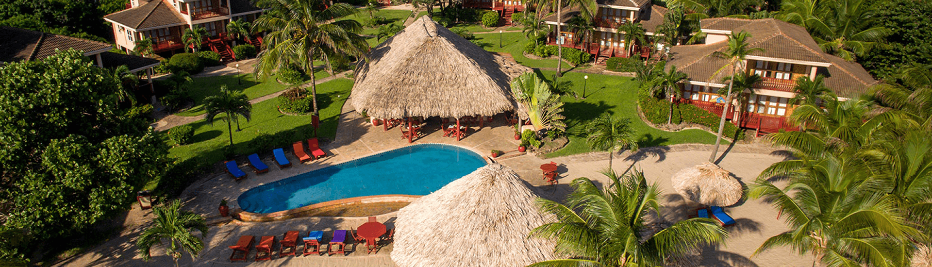 An aerial view of a tropical resort with a swimming pool surrounded by thatched-roof buildings and palm trees. Lounge chairs and umbrellas are scattered around the pool area.