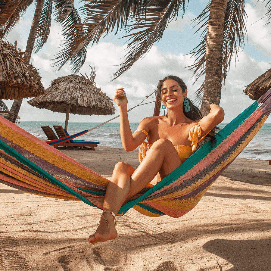 Woman in a bathing suit sitting in a multi-color hammock, with large green square earrings, with grass umbrellas, sand, ocean and palm trees in the background.