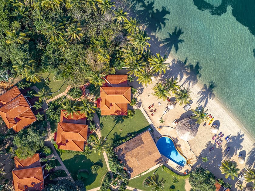 Aerial view of tropical resort with red-roofed villas, a pool, and a beach surrounded by palm trees.