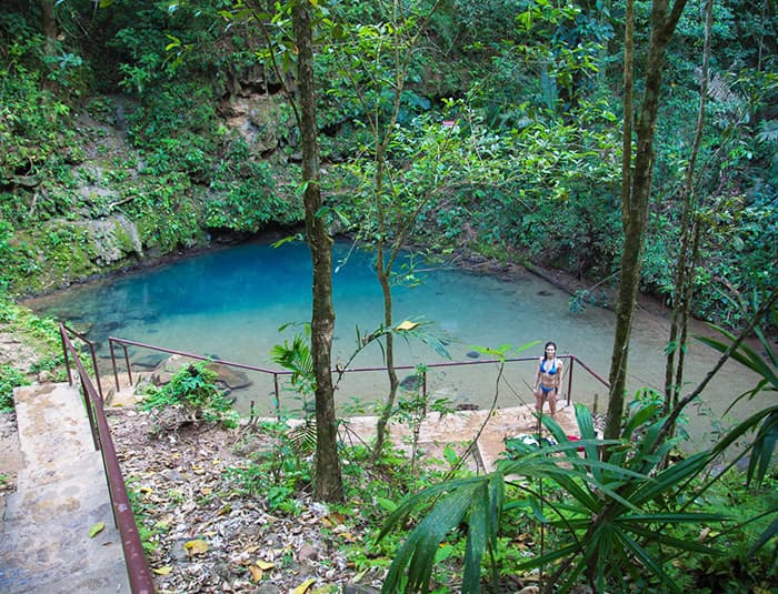 The image shows a beautiful, clear blue pool surrounded by lush greenery. A woman is standing in the water, and there are stairs leading down to the pool. It looks like a peaceful and refreshing spot to swim.